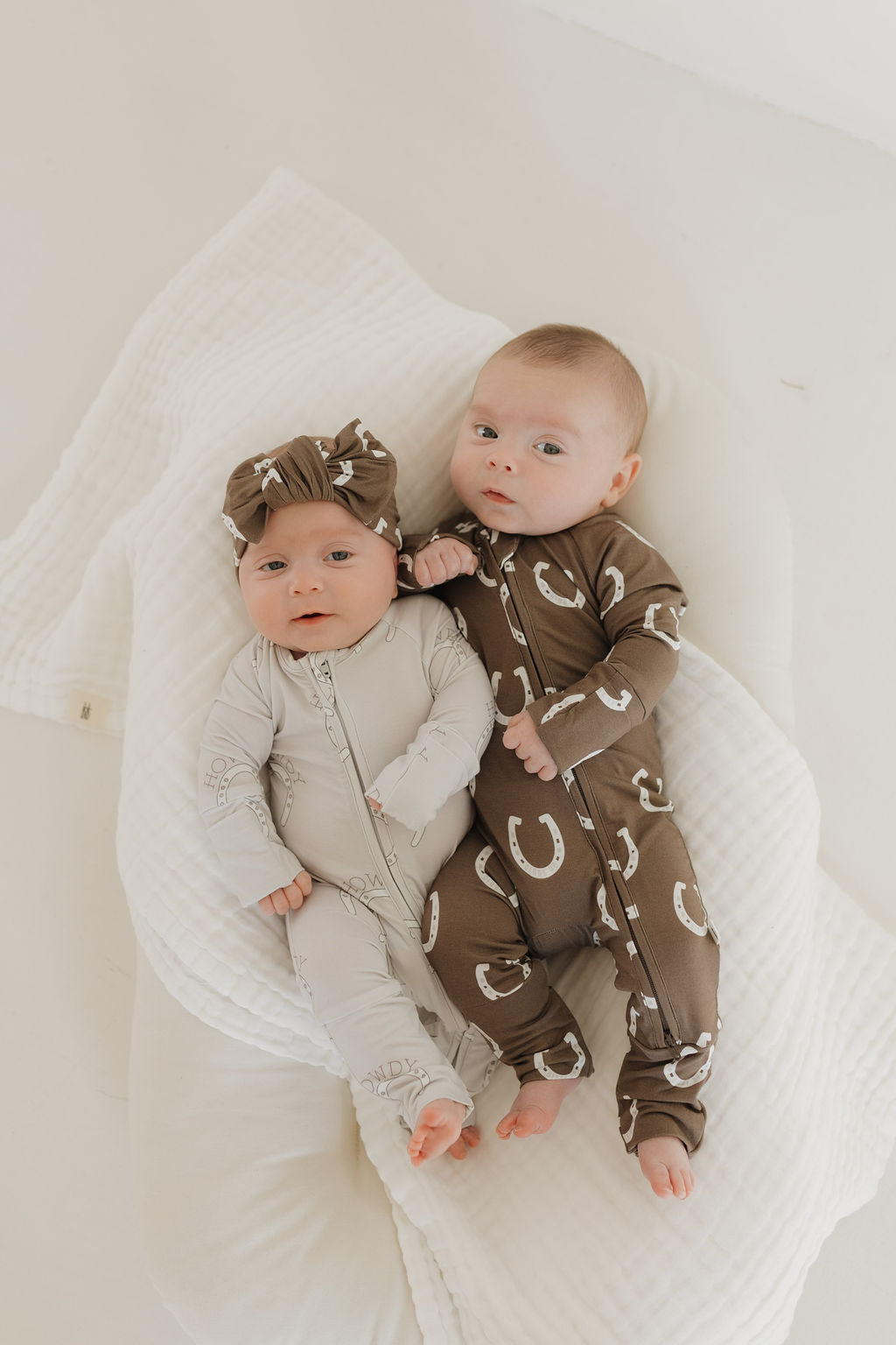 Two babies lie on a soft, white surface. One wears a light gray onesie with a matching headband, the other in patterned brown pajamas from forever french baby's Bamboo Zip Pajamas | Howdy line, ensuring comfort and breathability as they gaze up serenely.