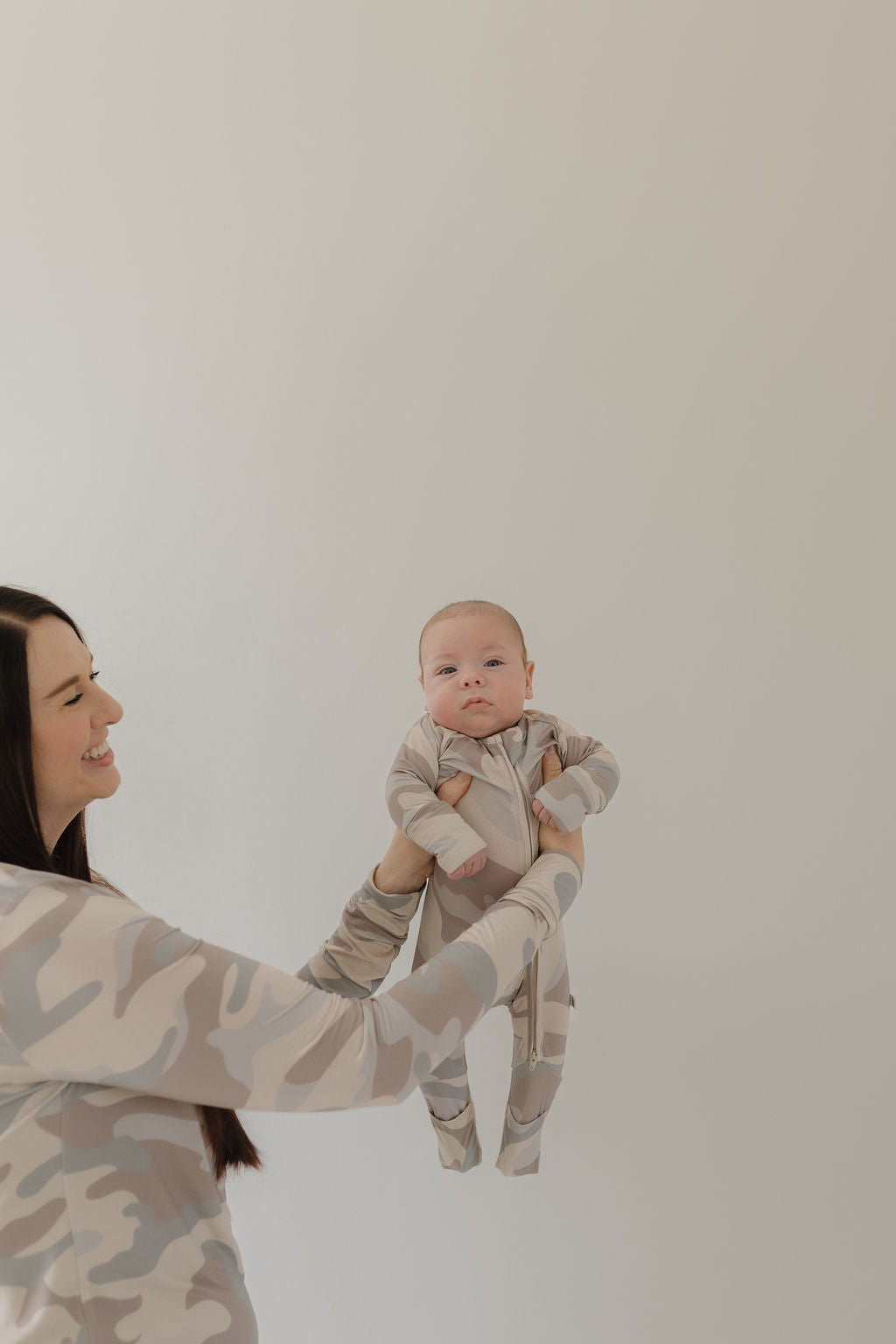 A person with long dark hair holds a baby, both in matching "Bamboo Zip Pajamas | Camo" from forever french baby. Against an off-white wall, the baby gazes at the camera, highlighting comfort and style.