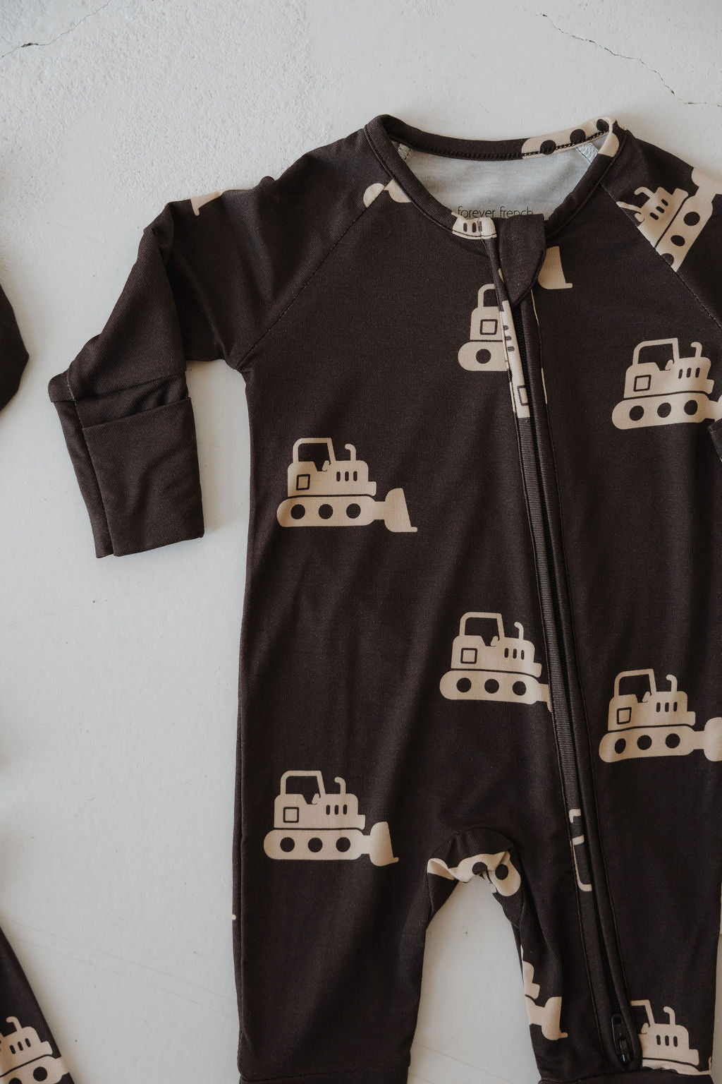 The forever french baby Bamboo Zip Pajamas | Bulldozer feature a dark brown, long sleeve onesie with a beige bulldozer pattern and zipper, made from breathable fabric and shown laid flat on a light surface.