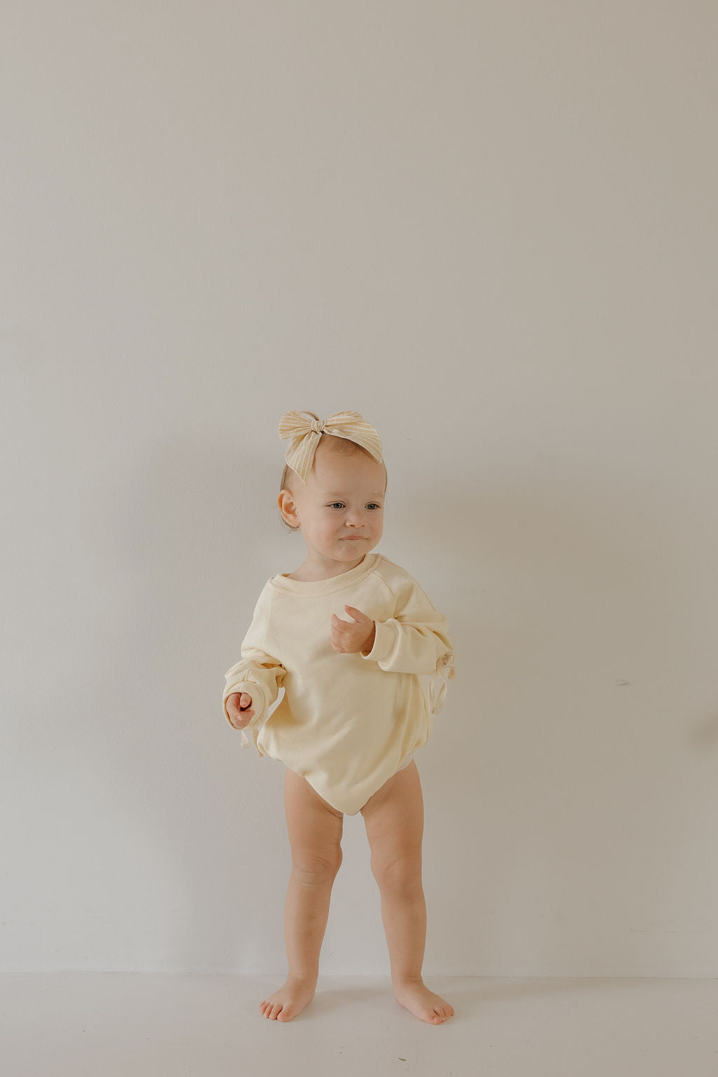 A toddler models the forever french baby Sweatshirt Romper | Buttercream Bows with a matching bow headband, barefoot by a light wall—ideal for cozy newborn wear or family matching outfits.