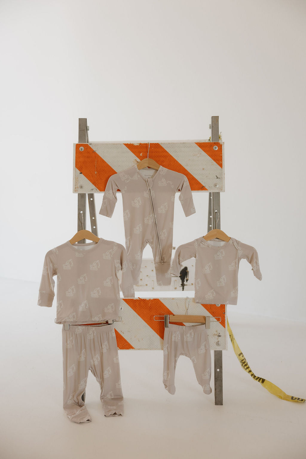 Four pieces from forever french baby's Sleepy Time Set | Excavator, in beige with white animal prints, hang on a construction barrier with orange and white stripes. A step ladder is visible in the background. Hypo-allergenic and for babies/toddlers.