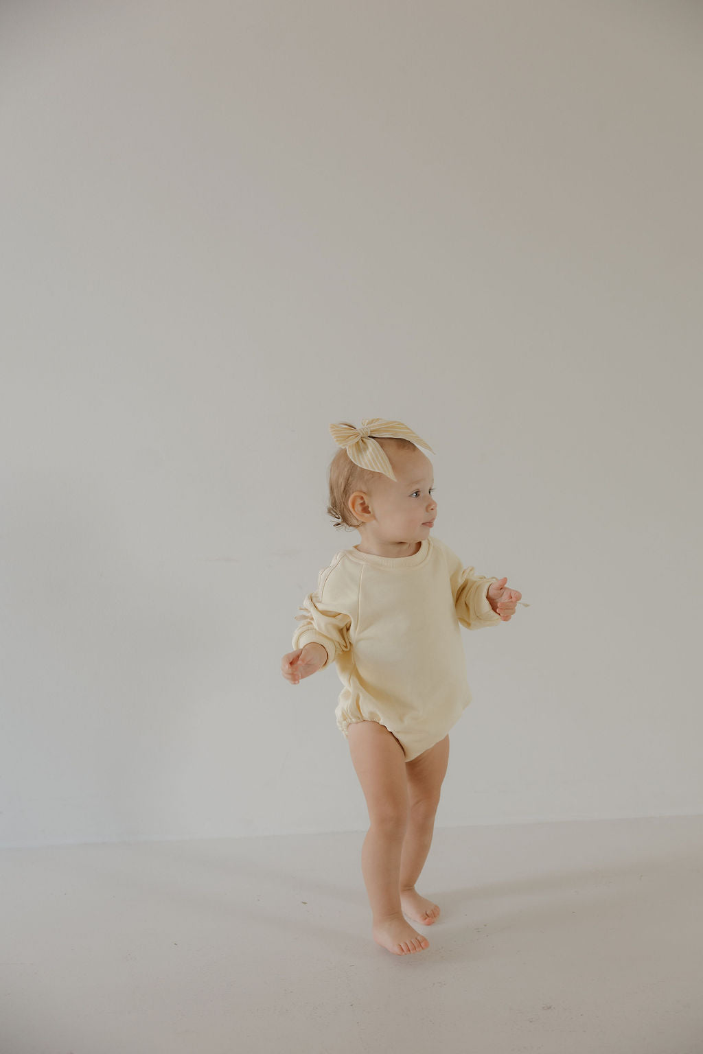 A toddler in the forever french baby Sweatshirt Romper | Buttercream Bows with a matching headband stands barefoot on a light floor, gazing to the side against a plain white background.