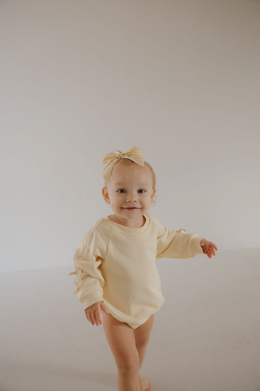 A smiling baby wears the forever french baby Sweatshirt Romper | Buttercream Bows, with a matching bow headband, standing barefoot on a light floor against a plain background.