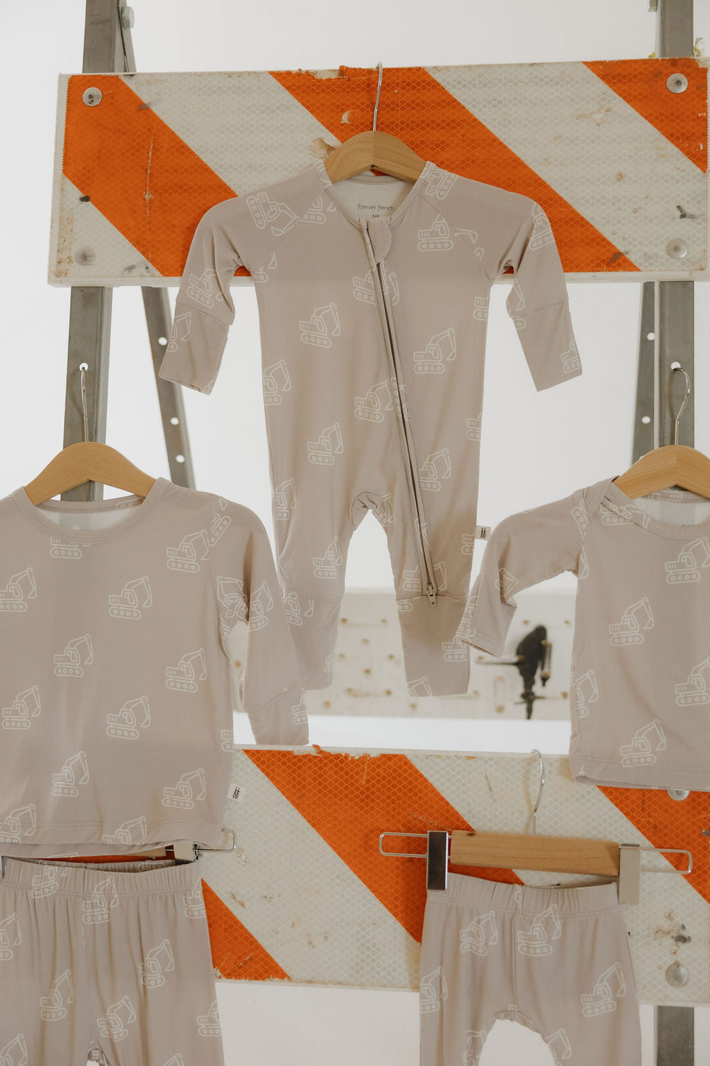 Three beige "Bamboo Zip Pajamas | Excavator" by forever french baby, featuring white construction vehicles, hang in front of an orange and white striped roadwork sign and a metal ladder.