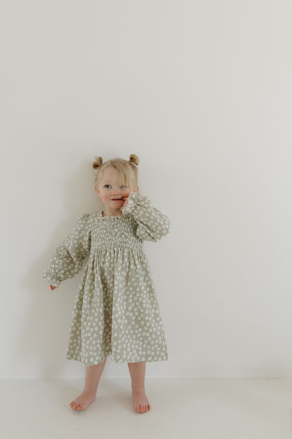 A young child with light hair in two buns stands against a white background, wearing the forever french baby "Child Dress | French Gray Floral." They look towards the camera with a relaxed expression, one hand touching their face.