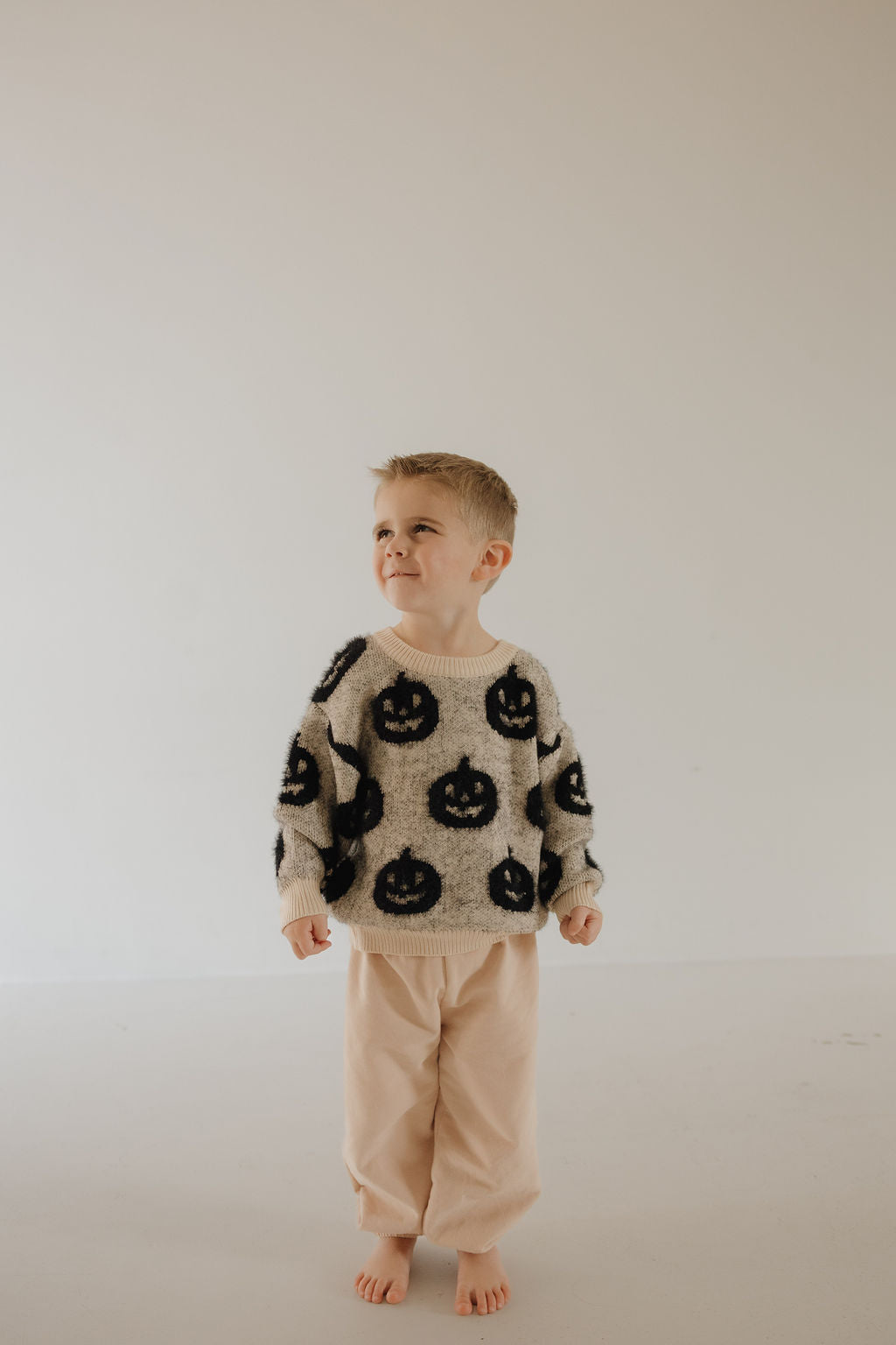 A young boy stands barefoot on a light floor in the forever french baby Cozy Sweater | Halloween, featuring black jack-o’-lantern faces. He smiles gently, looking to the side against a simple, bright background.