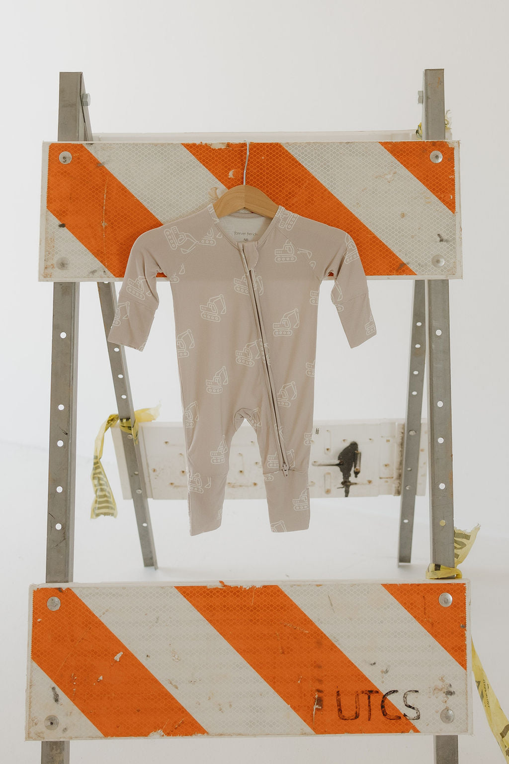 The Bamboo Zip Pajamas | Excavator by forever french baby, crafted from breathable bamboo with white animal prints, is displayed on a wooden hanger against a plain white background, hanging from a metal barricade with orange and white stripes.