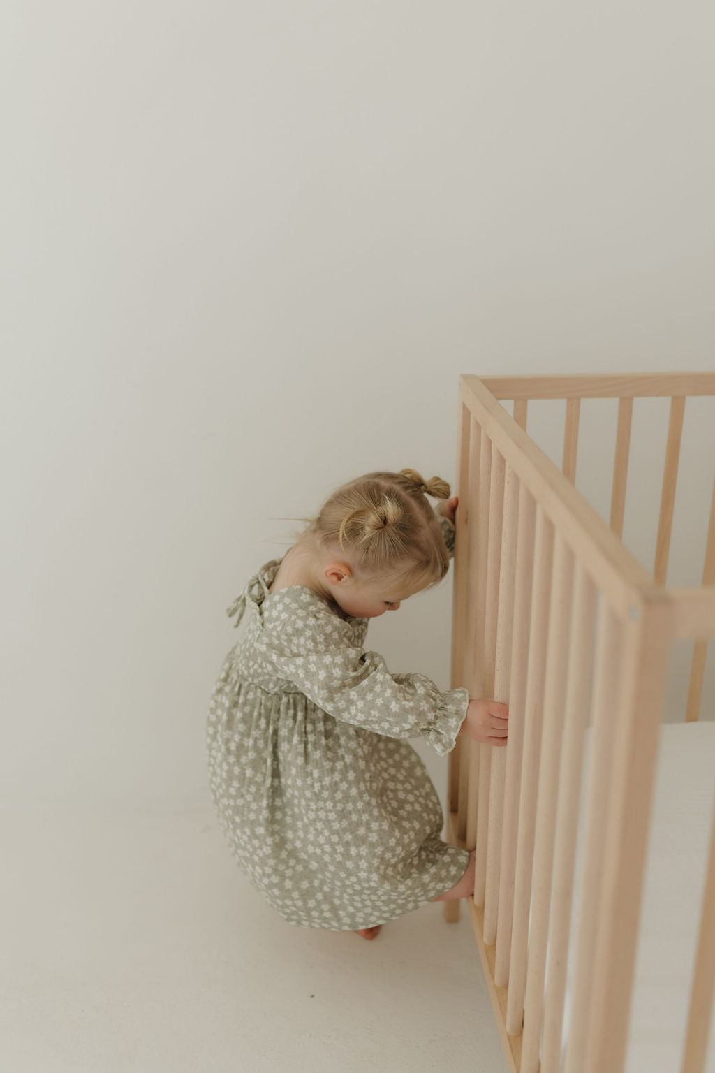 A toddler with light brown pigtails, dressed in the "French Gray Floral" cotton long sleeve dress by forever french baby, stands beside a wooden crib. She grasps the crib railing with both hands, slightly leaning forward against a neutral background.