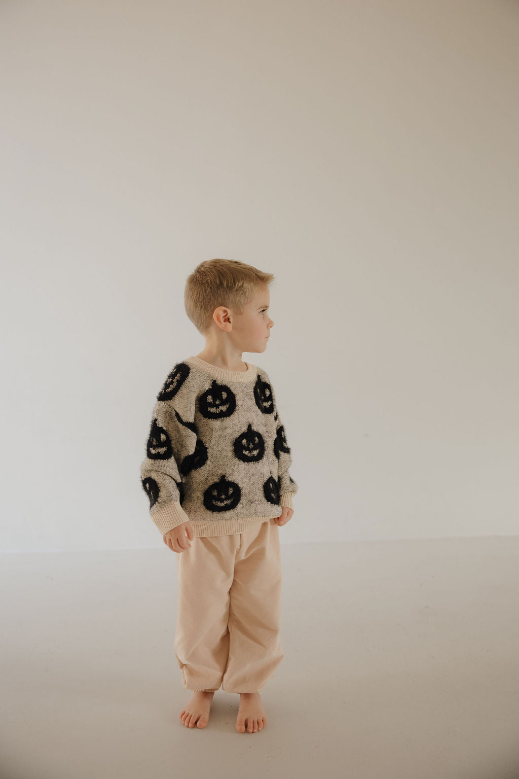 A young child with short blond hair stands barefoot on a light floor, wearing beige pants and the Cozy Sweater | Halloween by forever french baby—an adorable seasonal essential—looking to the side against a plain, light background.