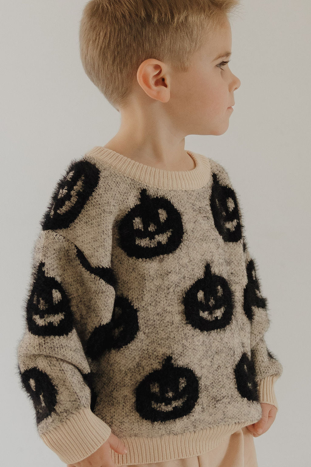 A young child with short blond hair models the forever french baby Cozy Sweater | Halloween, a beige knit with black jack-o'-lantern faces, standing in profile against a plain light background.