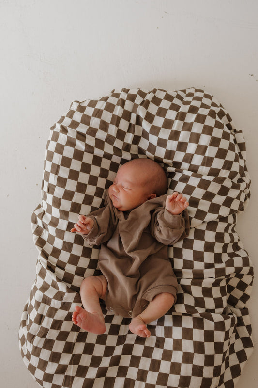 A newborn sleeps peacefully in the forever french baby Sweatshirt Romper | Hot Cocoa, resting on a brown-and-white checkered blanket with a white background.