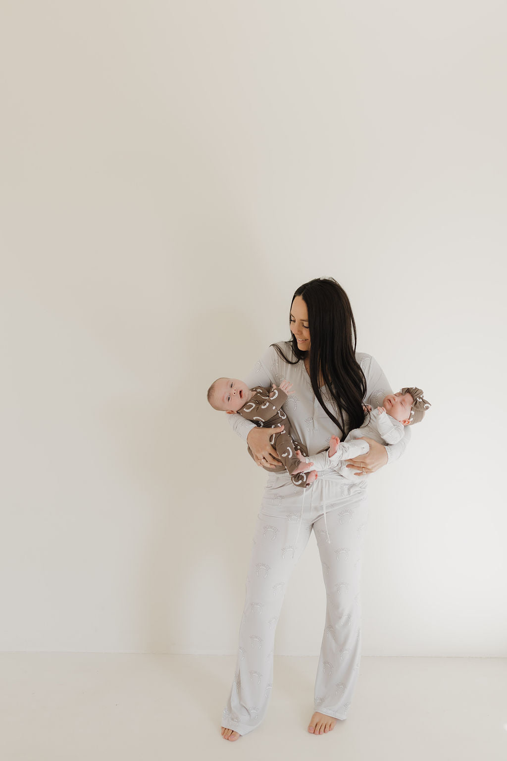 A woman with long dark hair cradles two babies against a light background. She wears a light bamboo pajama set from forever french baby, while both babies are in matching brown onesies with animal ear hats.