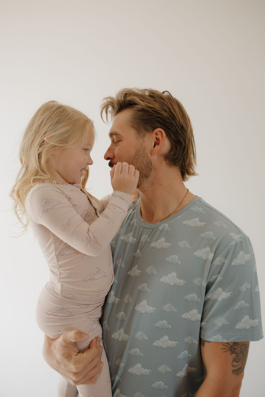 A man holds a smiling young girl as they touch noses, both wearing Kendy 3's Bamboo Two Piece Pajamas | Ruby. Their matching bamboo pajamas feature cloud patterns and stand out against a plain, light background.