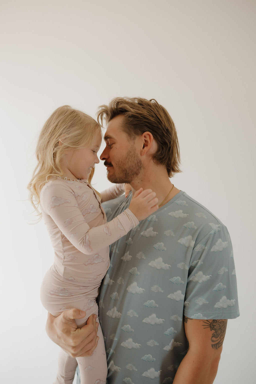A man wears the Kendy 3 Men's Short Sleeve Bamboo Pajama (Duke) and holds a smiling young girl as they touch noses, both in light-colored, cloud-patterned, breathable bamboo sets against a plain white background.