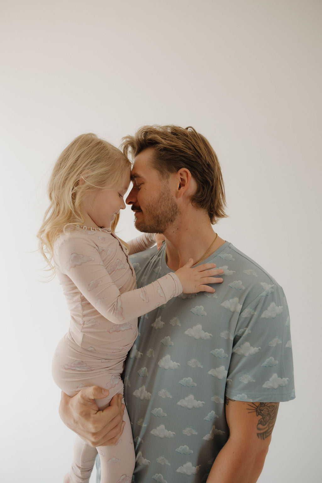 A man holds a young girl in his arms as they smile and touch foreheads, both wearing matching Kendy 3 Men's Short Sleeve Bamboo Pajama | Duke sets with a cloud pattern, made from hypo-allergenic, breathable fabric, against a plain light background.