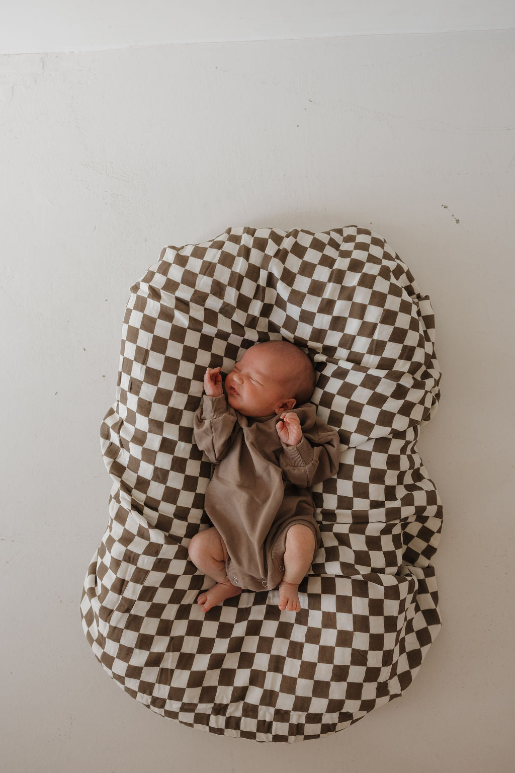 A newborn wearing the forever french baby Sweatshirt Romper | Hot Cocoa rests on a brown-and-white checkered cushion, set against a plain white background.