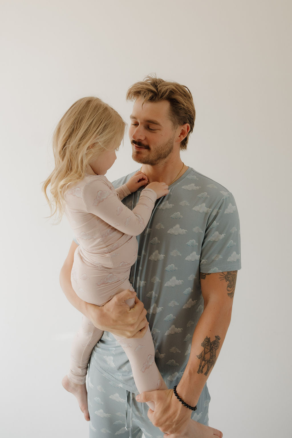 A man in Kendy 3’s Men’s Short Sleeve Bamboo Pajama | Duke holds a young girl in light pink pajamas, both smiling at each other against a plain background, enjoying the pajama's breathable and hypo-allergenic comfort.