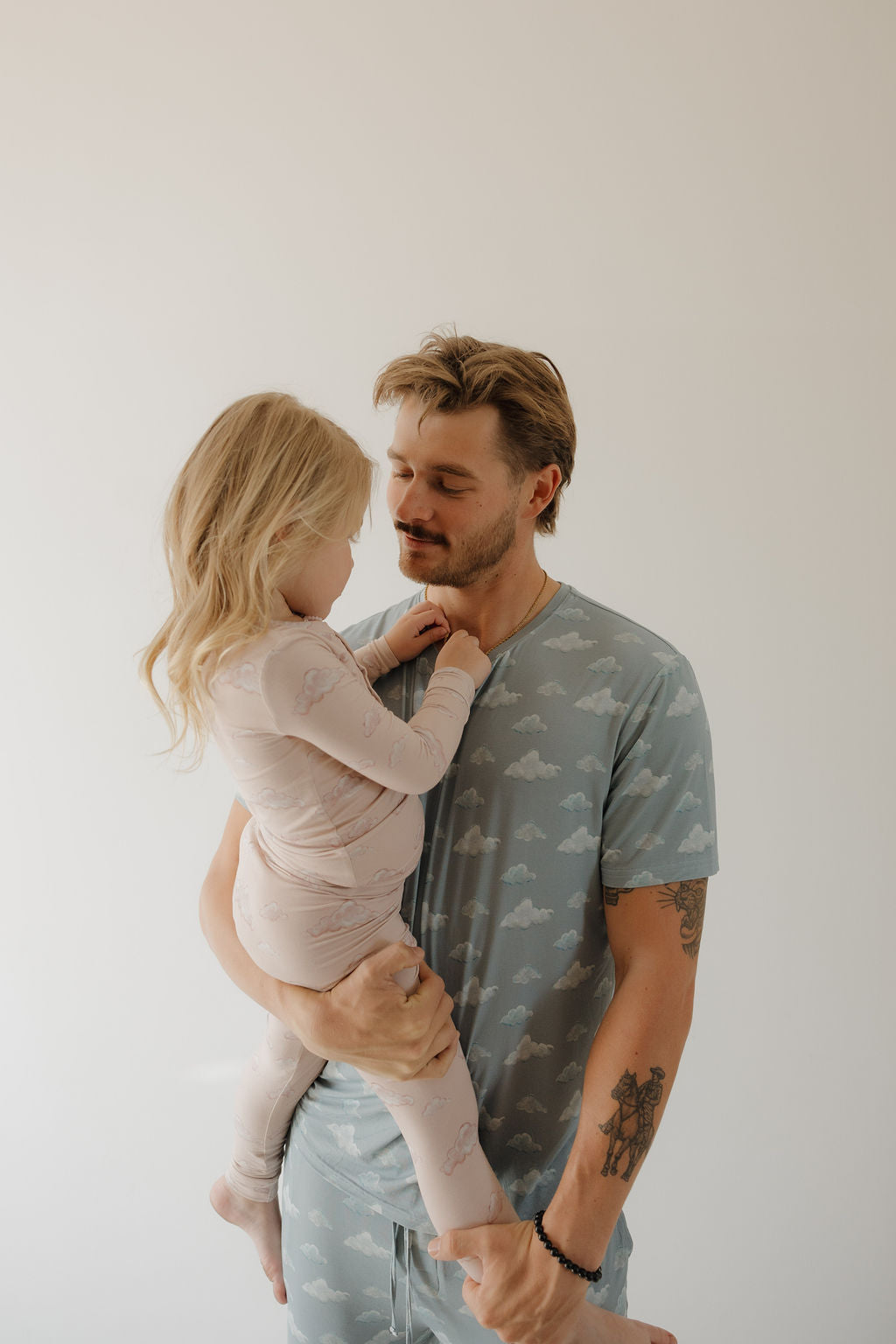 A man wears the Kendy 3 Men's Short Sleeve Bamboo Pajama | Duke in blue with a cloud pattern, holding a young girl in pink pajamas. They share an affectionate look against a light background, enjoying the breathable comfort.