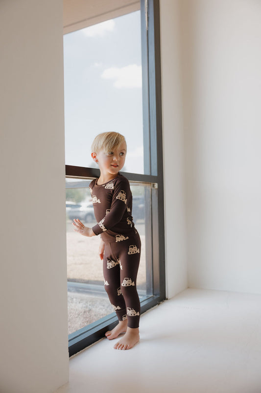 A young child wears the Child Sleepy Time Set | Bulldozer by forever french baby, standing by a large window and gazing outside. Sunlight fills the bright, minimalistic room with white walls.