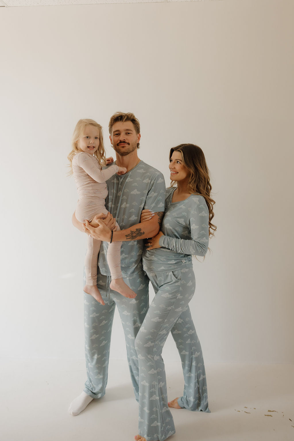 A man in Kendy 3 Bamboo Two Piece Pajamas | Ruby—blue set—holds a girl in pink pajamas, while a woman in matching blue bamboo pajamas smiles beside them, hand on her belly, against a plain light background.