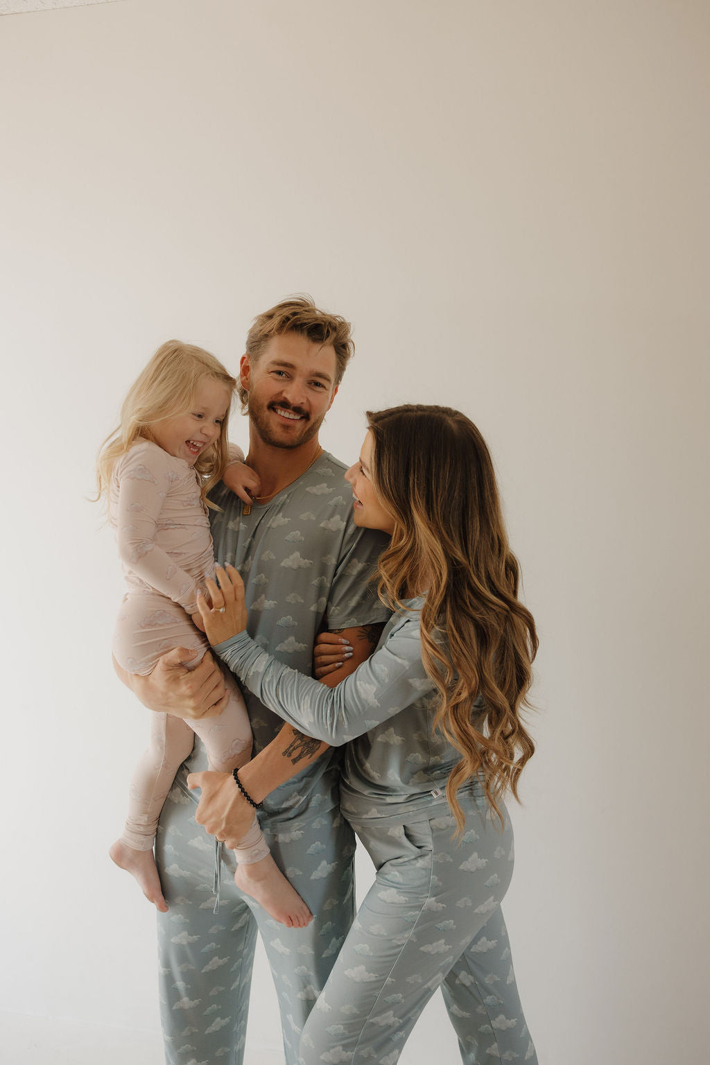 A smiling woman wears Kendy 3’s Women's Bamboo Pajama | Duke in light blue with cloud patterns, standing close to a man as he holds a laughing girl in pink pajamas. All look happy together against a plain light background.