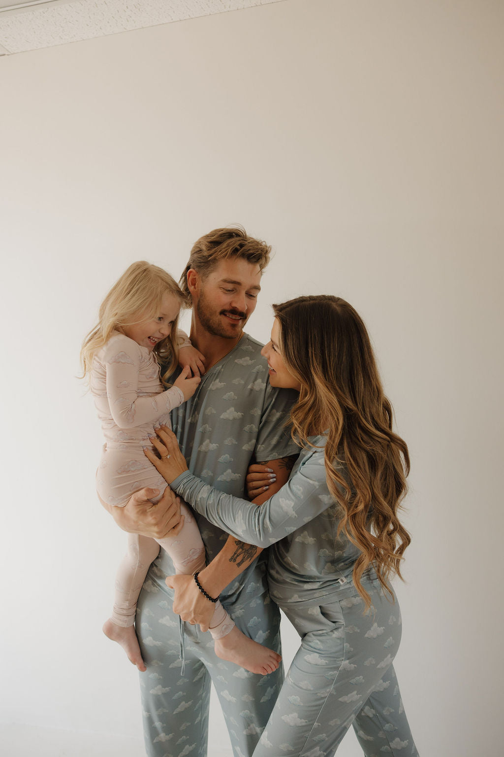A man and woman in matching blue Kendy 3 Women's Bamboo Pajama | Duke stand close, smiling at each other, while the man holds a smiling young girl in light pink pajamas looking at the woman.