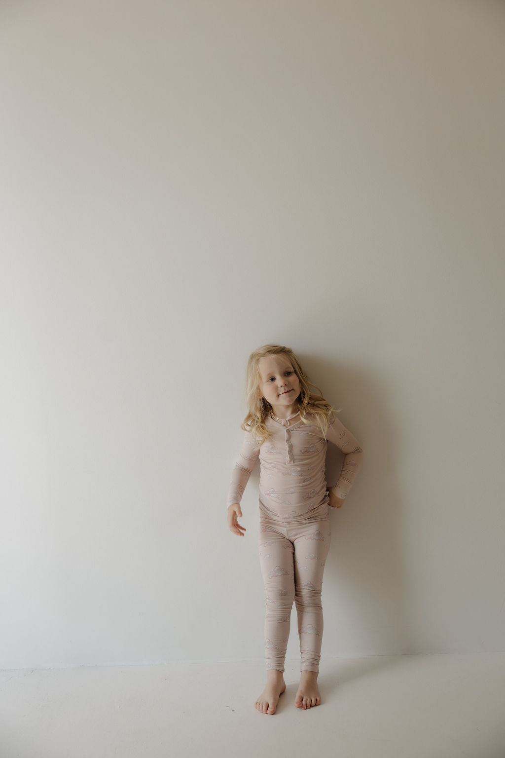 A young child with long blonde hair stands barefoot by an off-white wall, smiling gently and wearing Kendy 3’s Bamboo Two Piece Pajamas | Ruby—light beige, long-sleeve pajamas made from breathable bamboo fabric.
