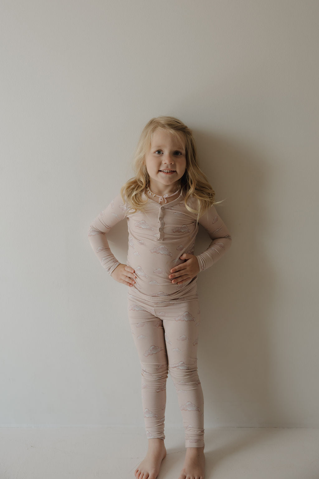 A young child with long blonde hair stands barefoot against a plain wall, smiling with hands on hips and wearing Kendy 3 Bamboo Two Piece Pajamas | Ruby, made from breathable bamboo fabric with a subtle pattern.