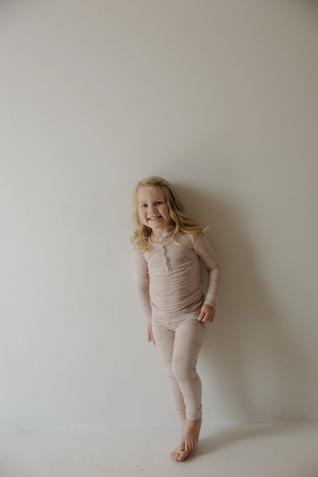 A young child with long blonde hair, barefoot, smiles at the camera while wearing Kendy 3’s Bamboo Two Piece Pajamas | Ruby in light pink, made from breathable fabric, standing against a plain off-white wall.