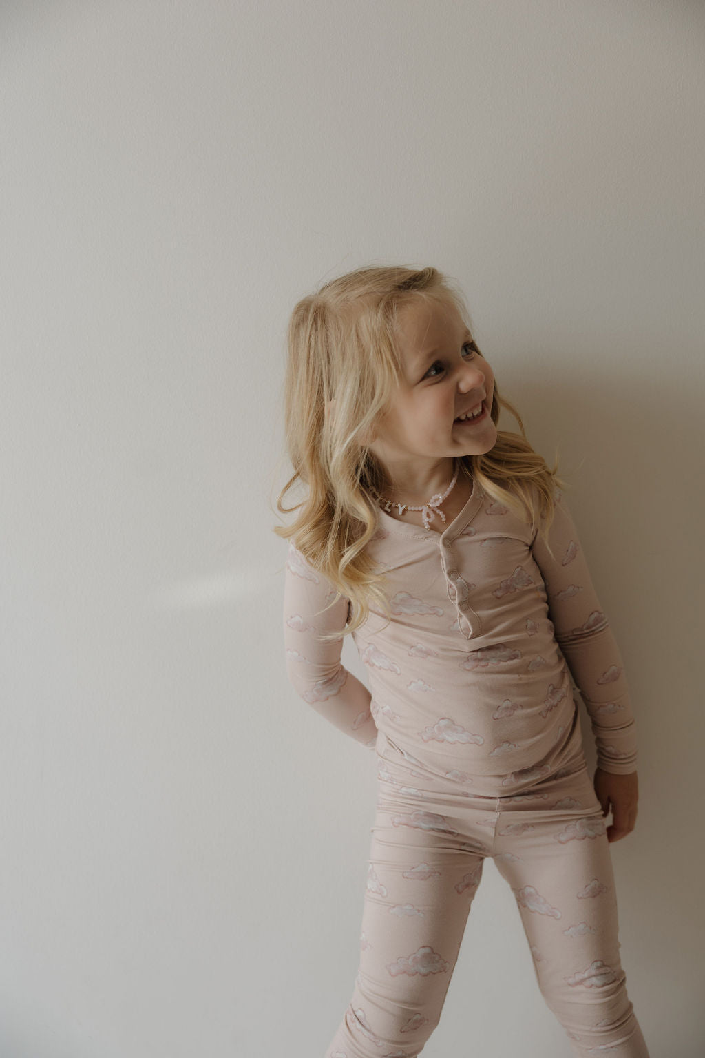 A young girl with long blonde hair smiles to the side, standing by a light wall in Kendy 3's Bamboo Two Piece Pajamas | Ruby—soft, breathable pajamas with a cloud pattern.