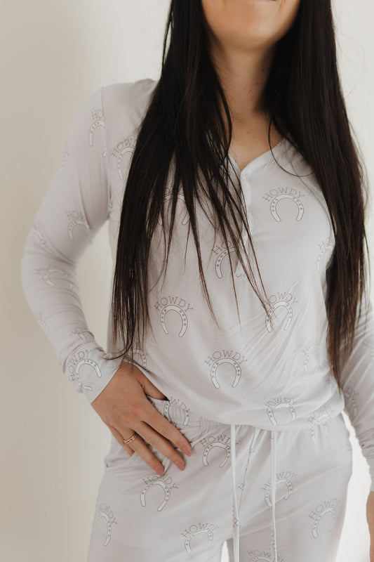 A woman with long dark hair models the "Women's Bamboo Pajama | Howdy" by forever french baby. The white, long-sleeved set features "Howdy" text and a horseshoe pattern. Made from hypoallergenic bamboo, these breathable pajamas blend style and comfort. The photo shows her from shoulders to mid-thigh, with one hand on her hip.