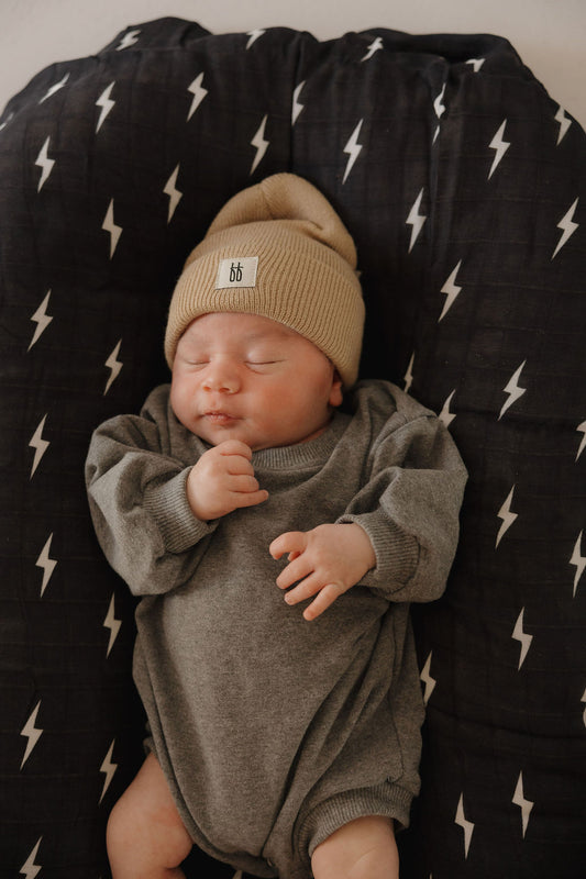 A newborn sleeps peacefully on a black cushion with white lightning bolts, wearing the forever french baby Sweatshirt Romper | Charcoal and a beige knit hat, one tiny hand resting near their face.