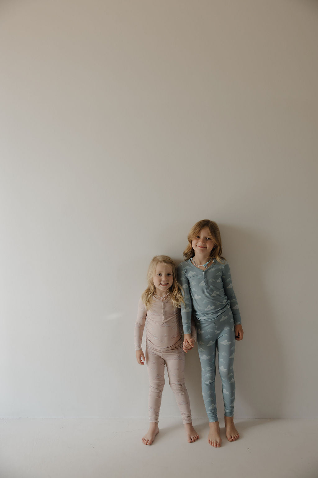 Two blonde children stand barefoot against a plain wall, smiling and holding hands in matching Kendy 3 Bamboo Two Piece Pajamas | Duke—long-sleeve pink and blue bamboo sleepwear that keeps them comfortable all night.