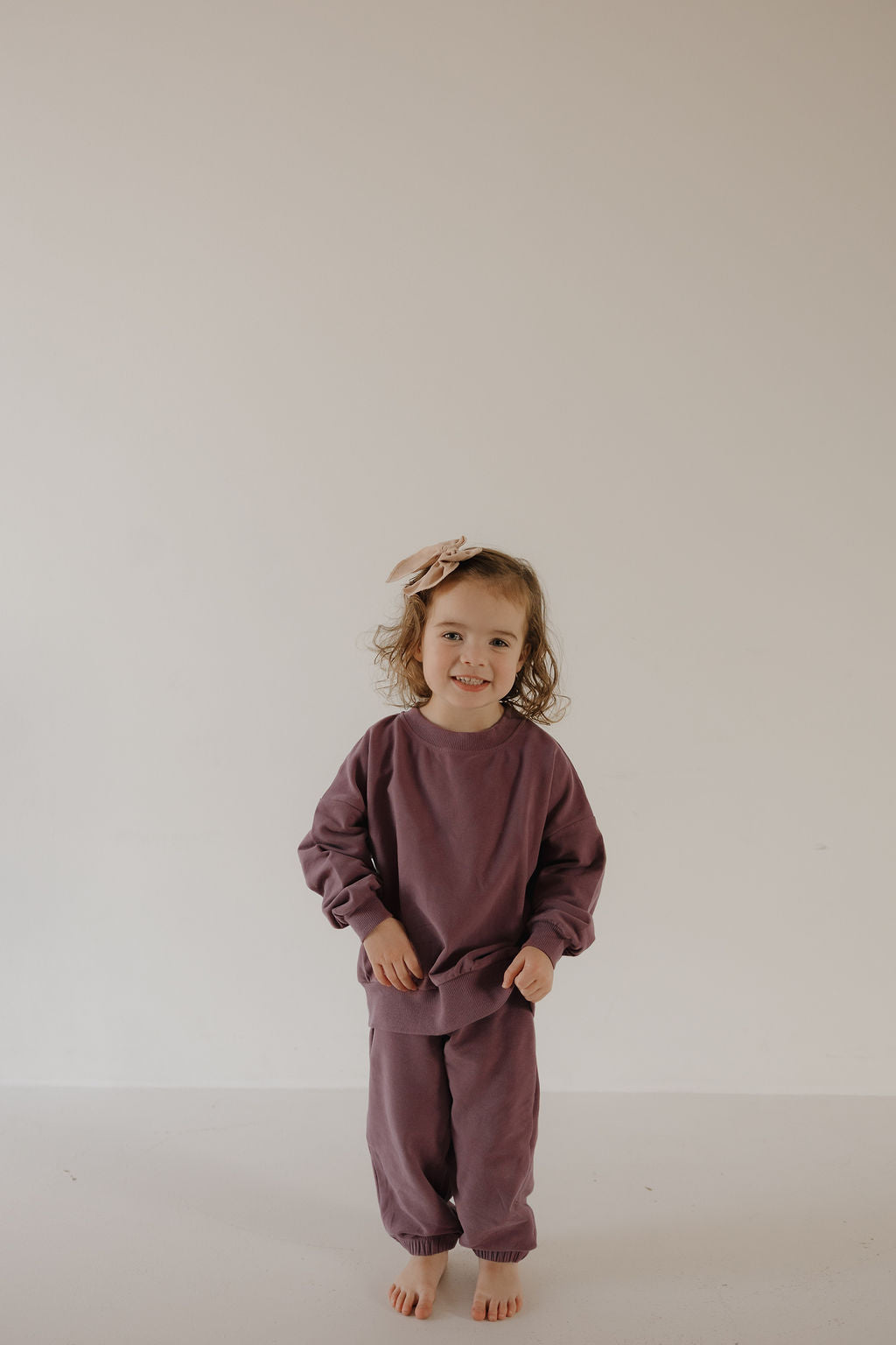 A young child with curly hair and a light bow smiles at the camera, wearing the forever french baby Child Sweat Set | Twilight. Barefoot against a plain background, she’s ready for a coordinated family look.