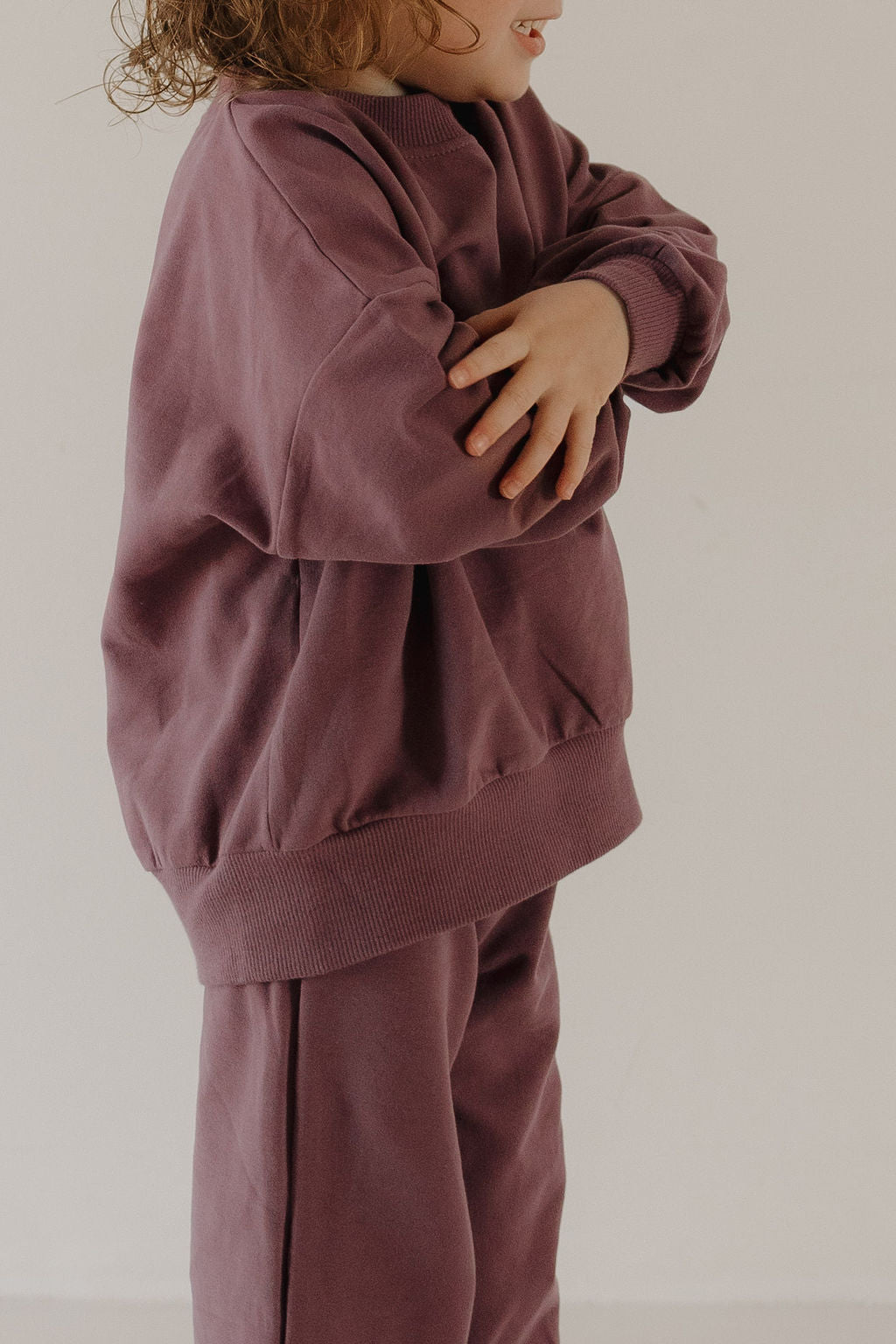 A young child in the forever french baby Child Sweat Set | Twilight stands with arms crossed against a neutral background, their face partially turned away and not fully visible.