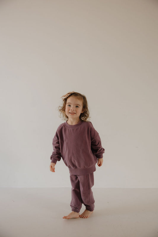A young child in the forever french baby Child Sweat Set | Twilight stands barefoot on a light floor against an off-white background, looking slightly to the side and smiling.