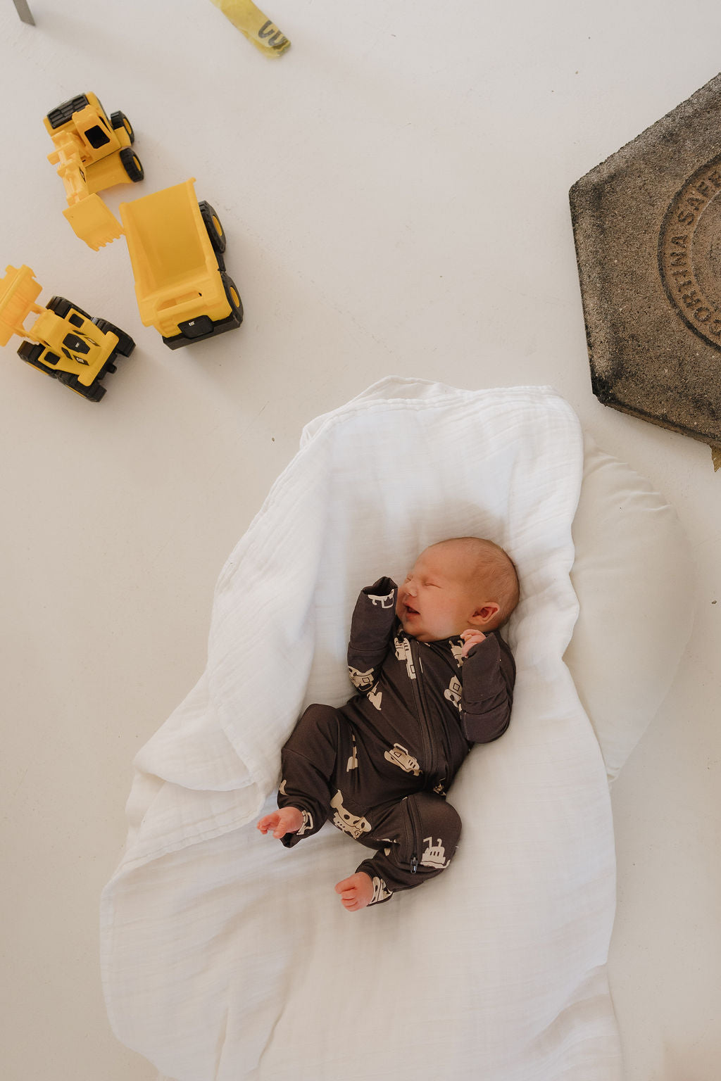 A newborn in forever french baby's Bamboo Zip Pajamas | Bulldozer sleeps on a white blanket. Yellow toy construction trucks and a rectangular textured mat are nearby, set against a light, minimal background.