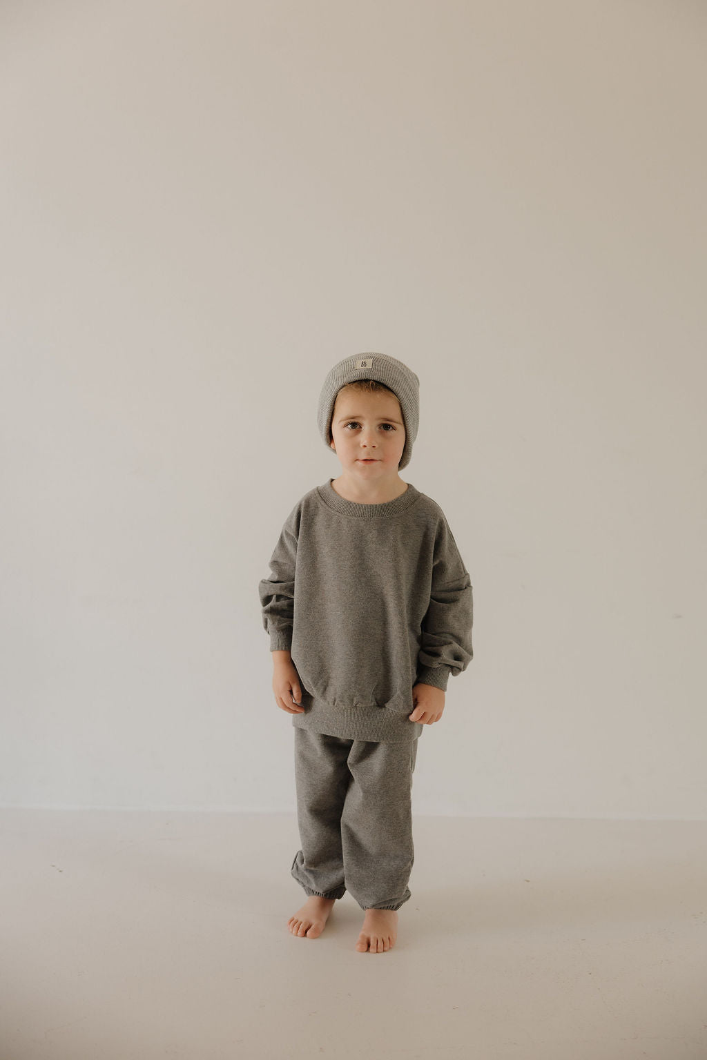 A young child wears the forever french baby Child Sweat Set in Charcoal—beanie, loose sweatshirt, and matching pants—against a light background for a relaxed, coordinated look. The child faces the camera with a neutral expression.