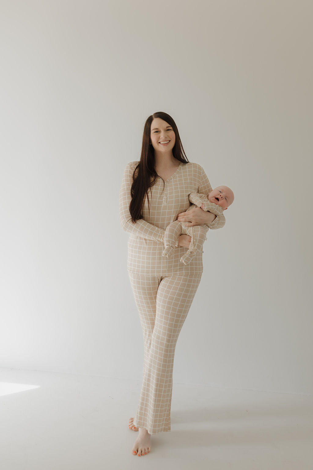 A woman smiles, cradling her baby in matching beige plaid outfits from forever french baby's Women's Bamboo Pajama | Grid, crafted from hypo-allergenic material for maximum comfort. A plain background enhances their serene connection.