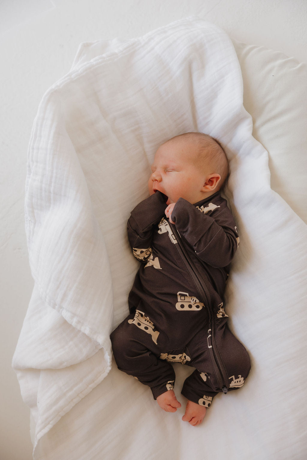 A newborn sleeps on a soft white blanket, wearing forever french baby Bamboo Zip Pajamas | Bulldozer—a dark brown onesie with light vehicle patterns—with one hand near their mouth and head turned slightly to the side.