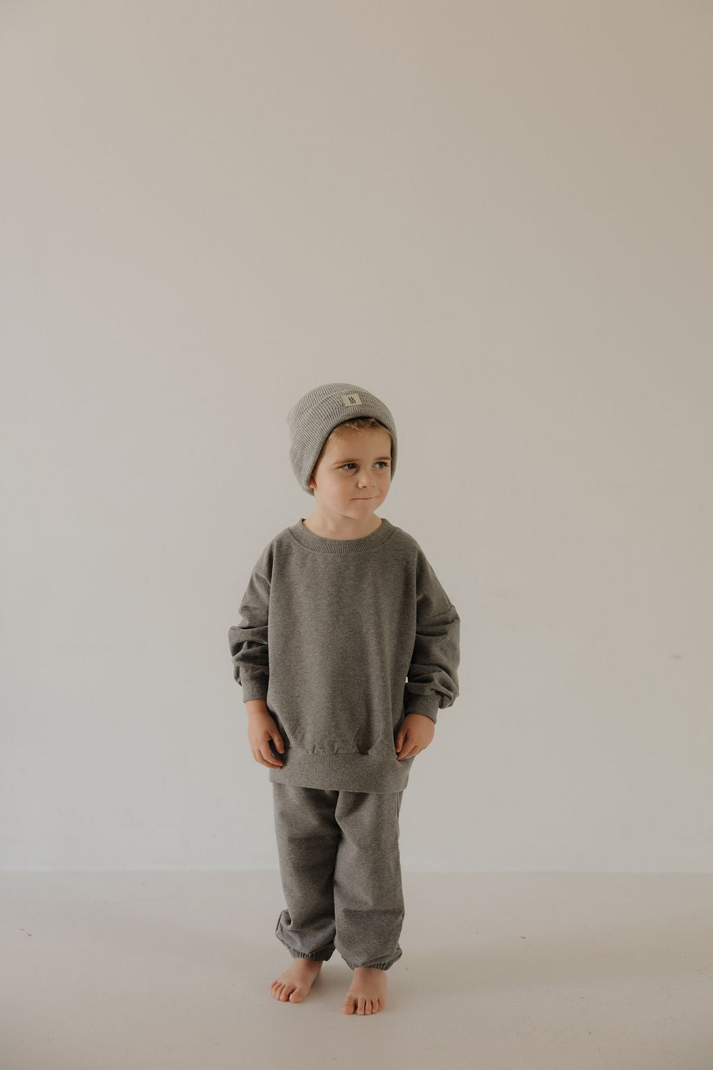 A young child stands barefoot on a light floor, wearing the forever french baby Child Sweat Set in Charcoal and a gray beanie, looking slightly to the side against a plain, light-colored background.