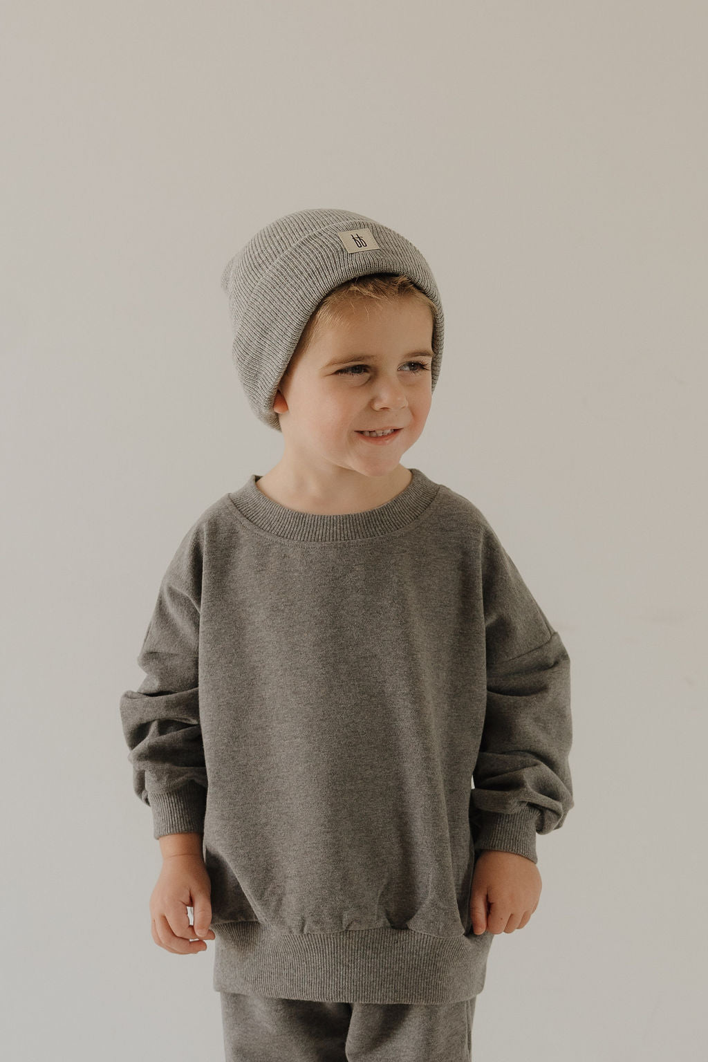 A young child in the forever french baby Child Sweat Set | Charcoal and a cozy gray knit beanie stands before a plain light background, smiling gently and glancing to the side.