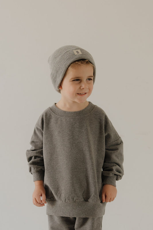 A young child in the forever french baby Child Sweat Set | Charcoal and a cozy gray knit beanie stands before a plain light background, smiling gently and glancing to the side.