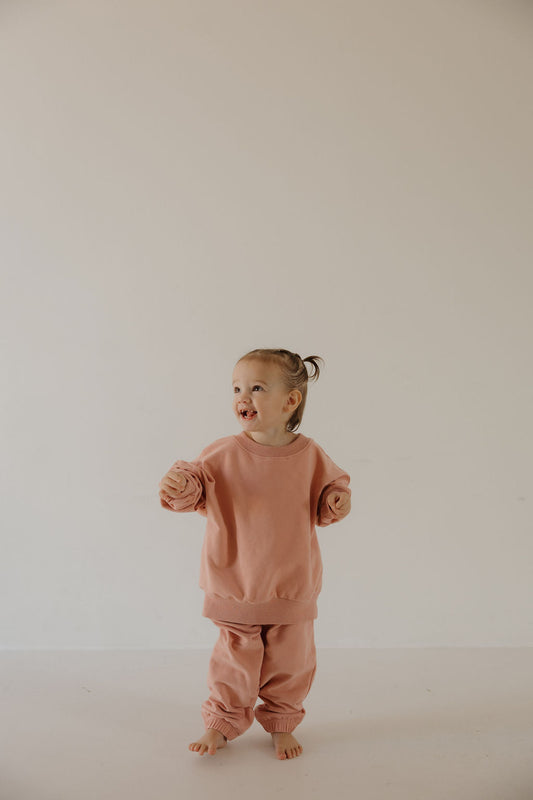 A young child with a small ponytail smiles against a light background, wearing the forever french baby Child Sweat Set in Rose—an oversized pink outfit perfect for everyday adventures.