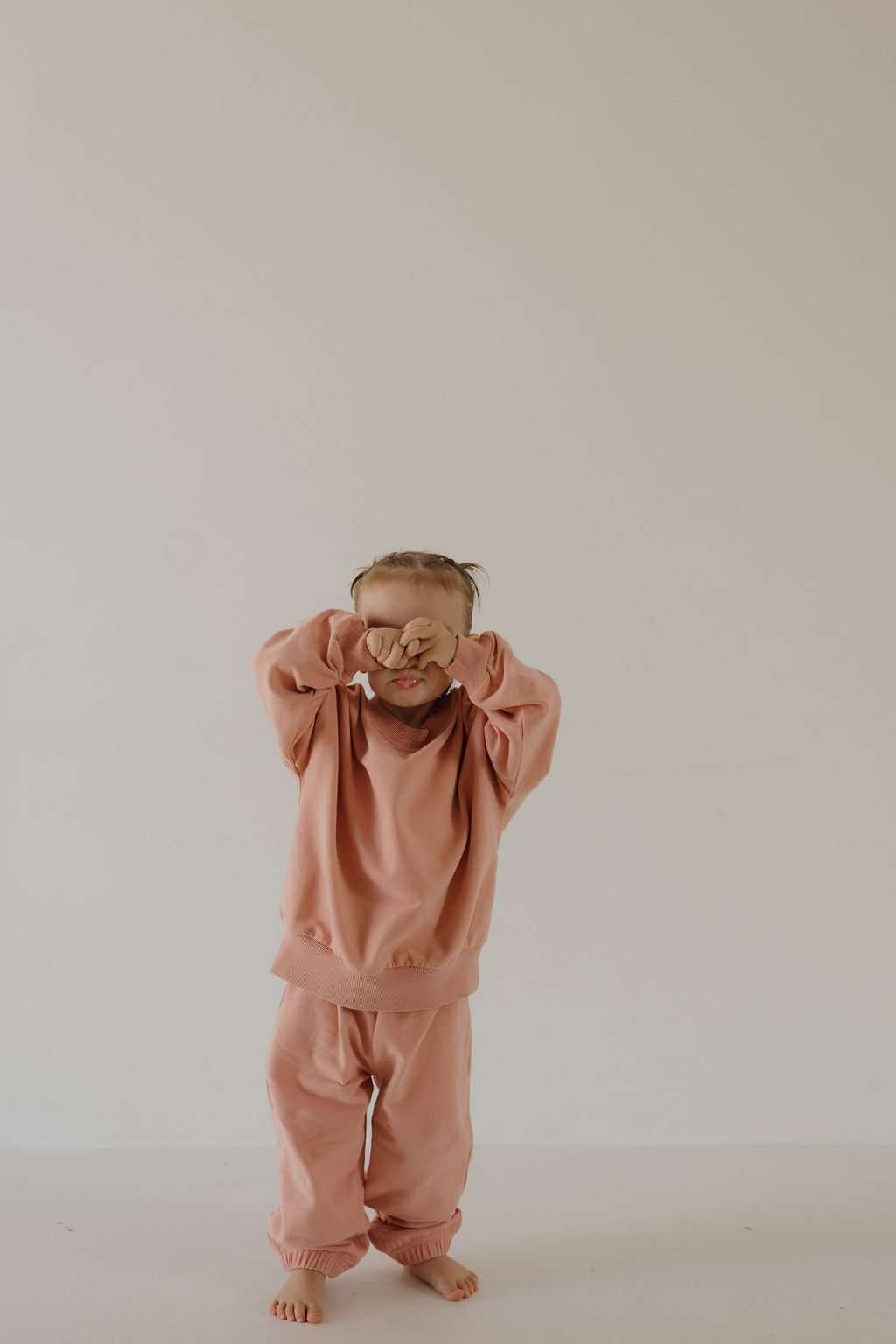 A young child wears the forever french baby Child Sweat Set | Rose, standing barefoot against a plain light background and covering their eyes with their hands, ready for everyday adventures.