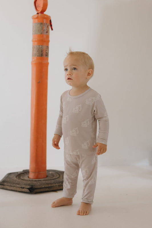 A toddler wears the forever french baby Child Sleepy Time Set | Excavator—light gray bamboo loungewear with white animal prints—standing barefoot on a light floor, with a red and silver pole behind and a plain, light-colored background.