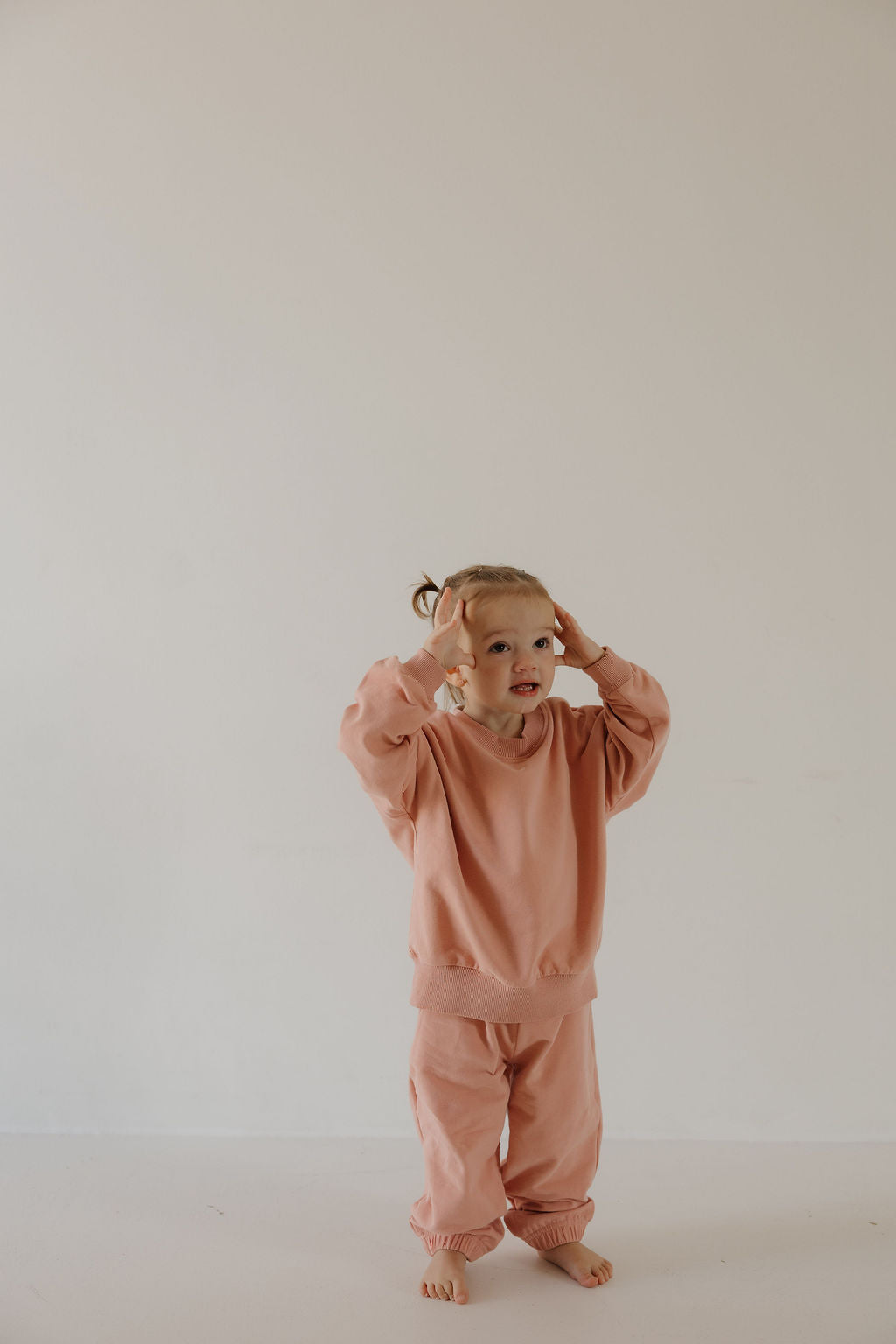 A young child with light hair stands barefoot against a white background, wearing the forever french baby Child Sweat Set | Rose, and touches their head with both hands while looking upward, ready for everyday adventures.