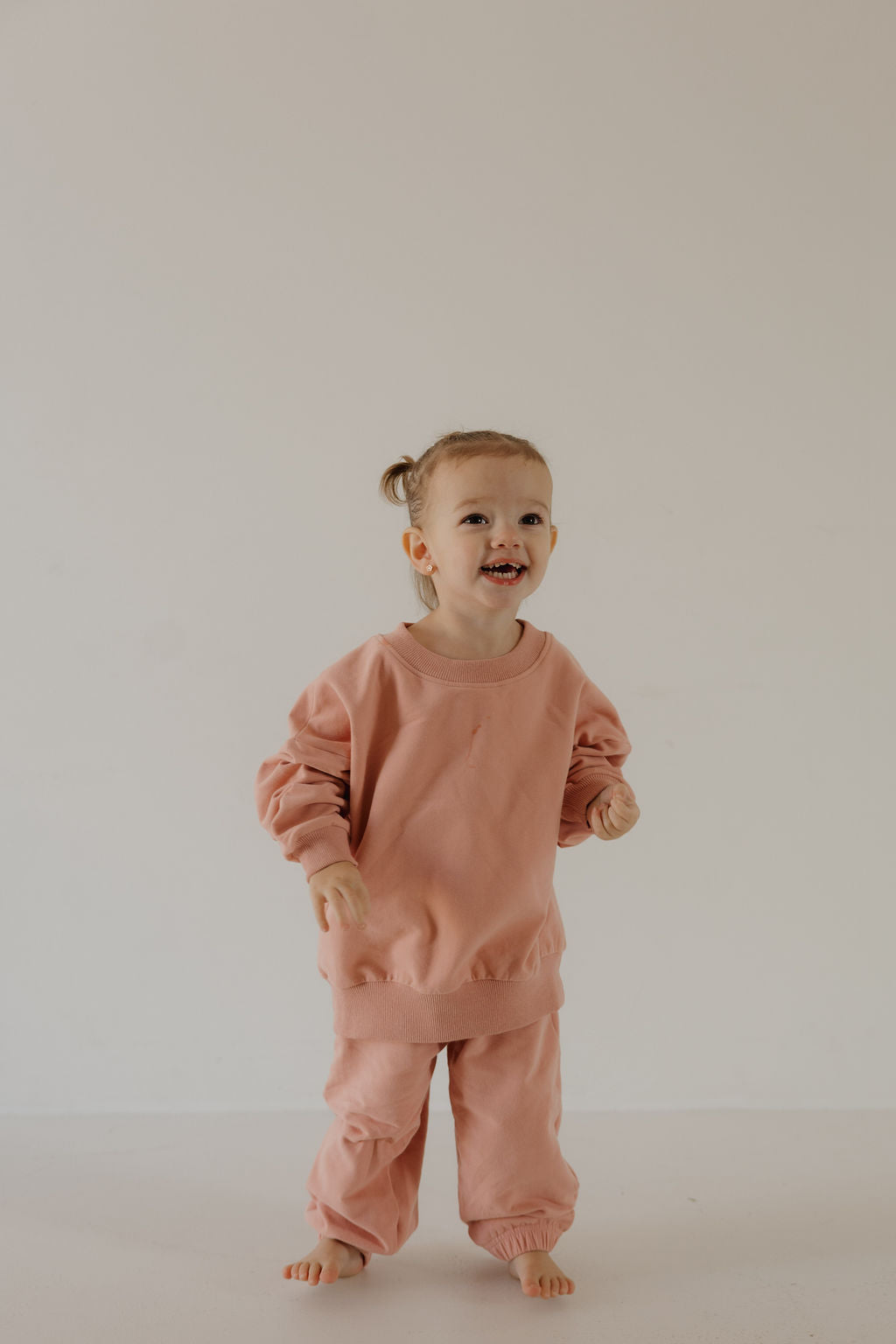 A young child with light hair in pigtails stands barefoot, smiling in a cozy, rose-colored Child Sweat Set by forever french baby—loose-fitting and perfect for everyday adventures against a plain, light background.