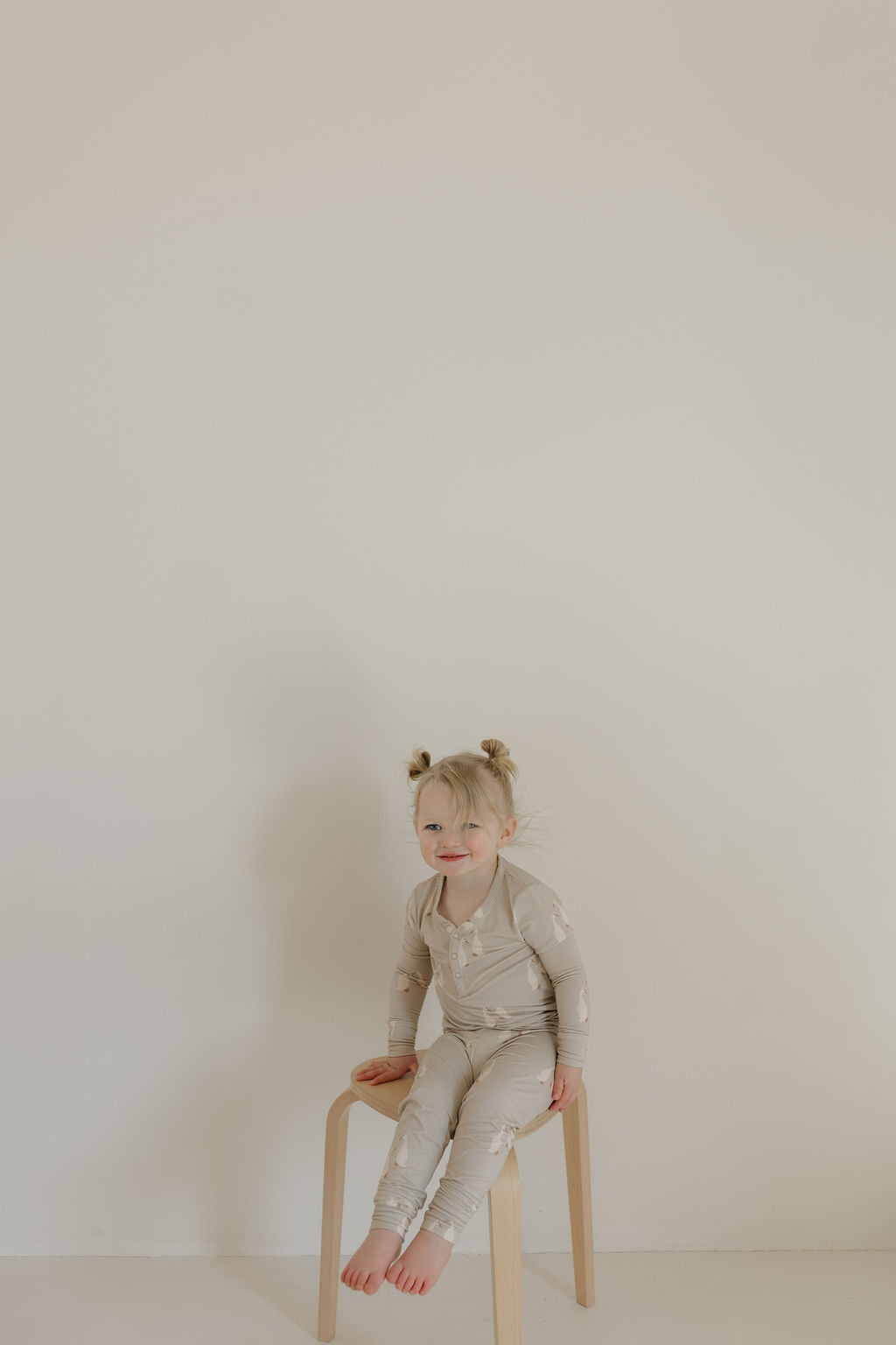 A smiling young child with light hair in two buns sits barefoot on a wooden stool against a white wall, dressed in breathable, hypo-allergenic Bamboo Two Piece Pajamas by forever french baby. The pajamas feature light-colored long sleeves with a subtle pattern.