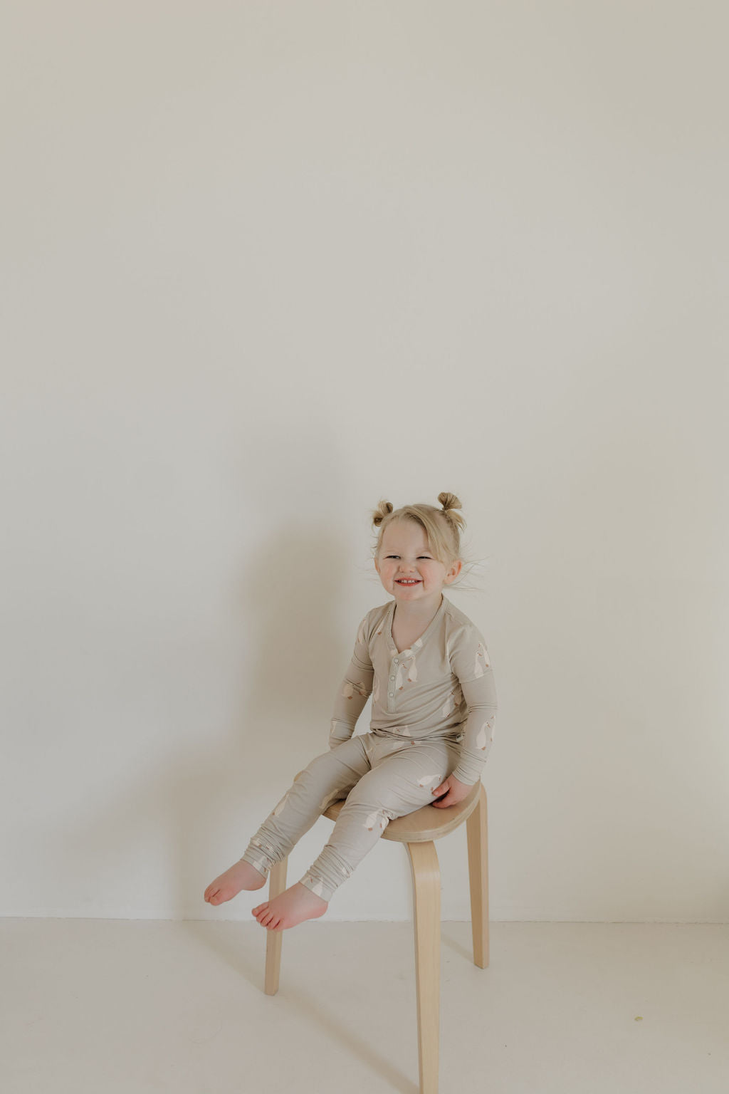 A toddler with blond hair in two tiny buns sits on a wooden stool, wearing full-length, patterned pajamas. These Bamboo Two Piece Pajamas by forever french baby | Silly Goose offer breathable comfort and style, while the cream-colored wall in the background enhances the minimalist setting.